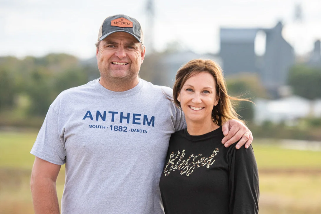 Taylor and Cassandra Sumption of Anthem Oats, smiling with their arms around each other in front of their South Dakota farm.