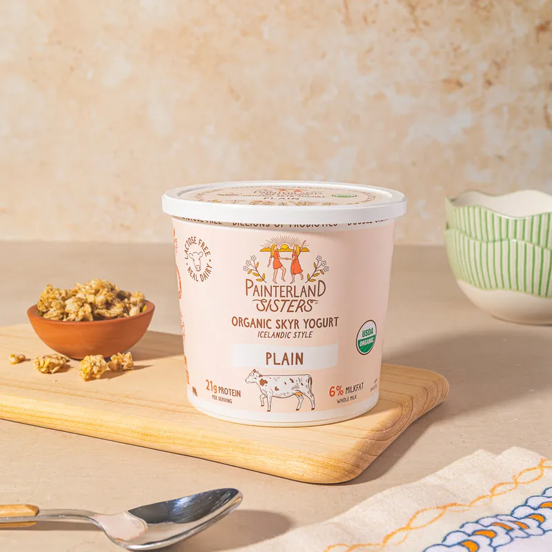 A tub of Painterland Sisters Organic Skyr Yogurt, Plain, on a wooden board next to a small bowl of granola.